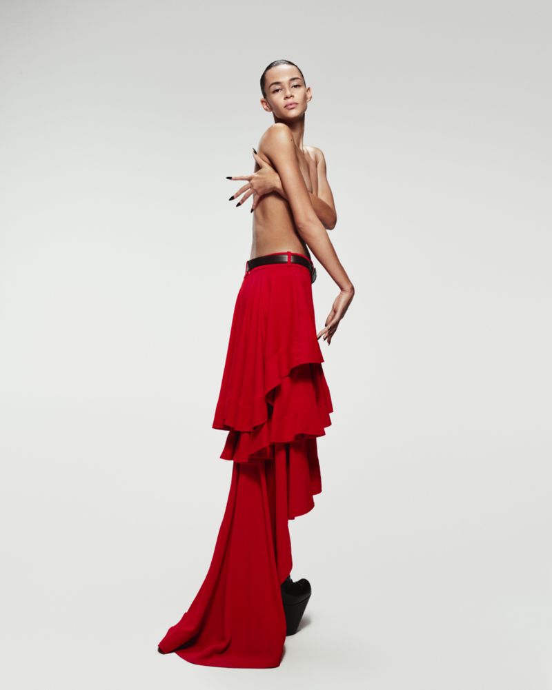 Adult posing with bare torso and a long, layered red skirt in a studio setting, hands raised. Fashion shot.
