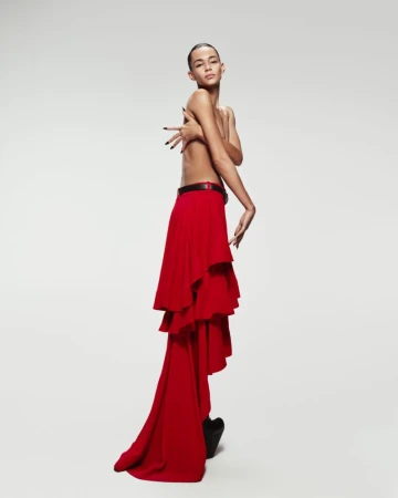 Adult posing with bare torso and a long, layered red skirt in a studio setting, hands raised. Fashion shot.