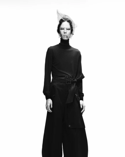 Monochrome studio shot of a slender adult fashion model in dark clothing with a veil, against a stark white backdrop, looking at the camera.