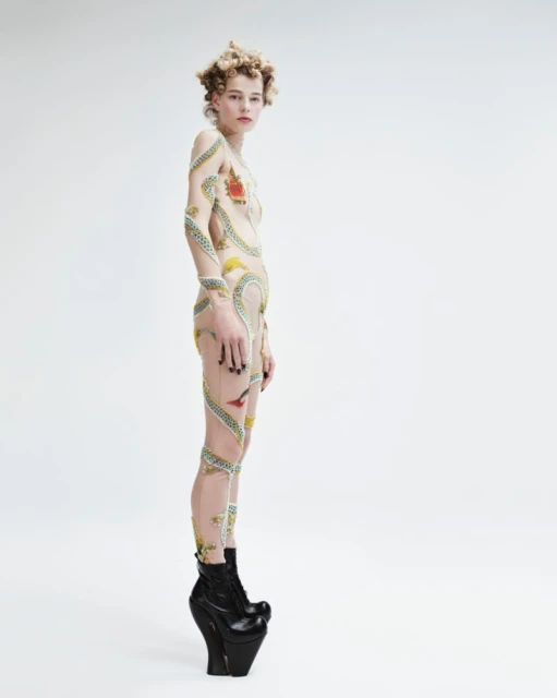 Full-length shot of person in decorative body suit and platform boots against a white backdrop, studio lighting, fashion shoot.