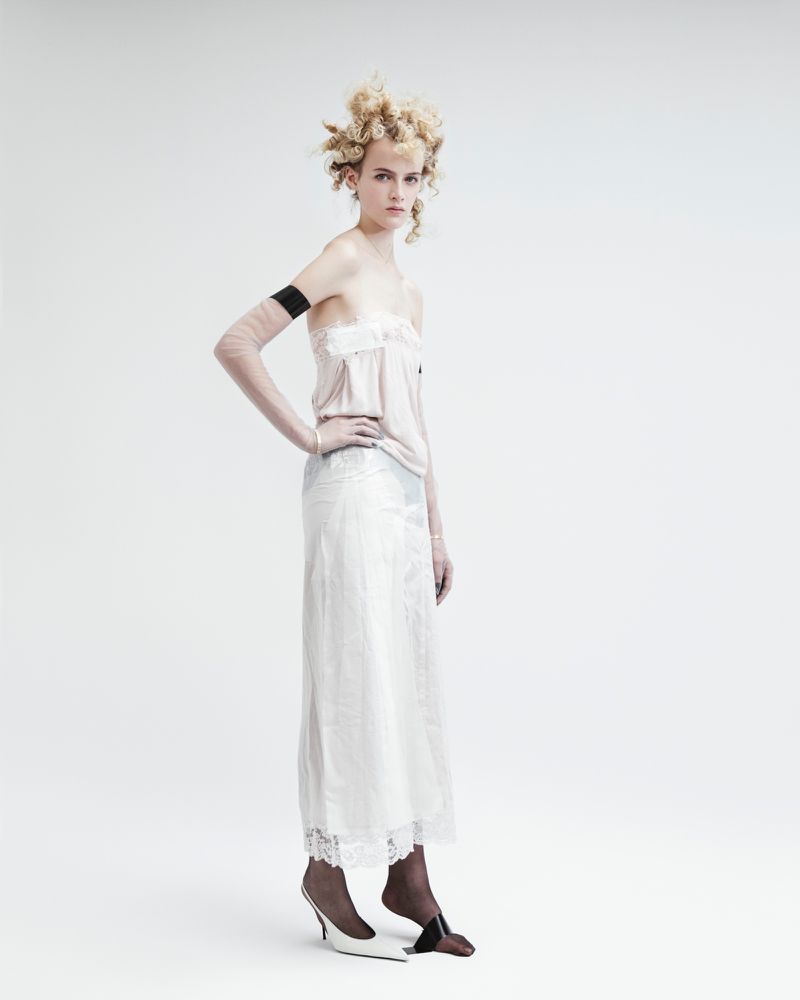 A woman poses in a light pink top and a long white skirt in a studio setting, highlighted by a clean, bright background.