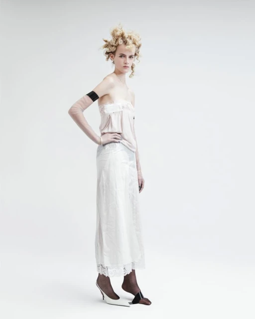 A woman poses in a light pink top and a long white skirt in a studio setting, highlighted by a clean, bright background.