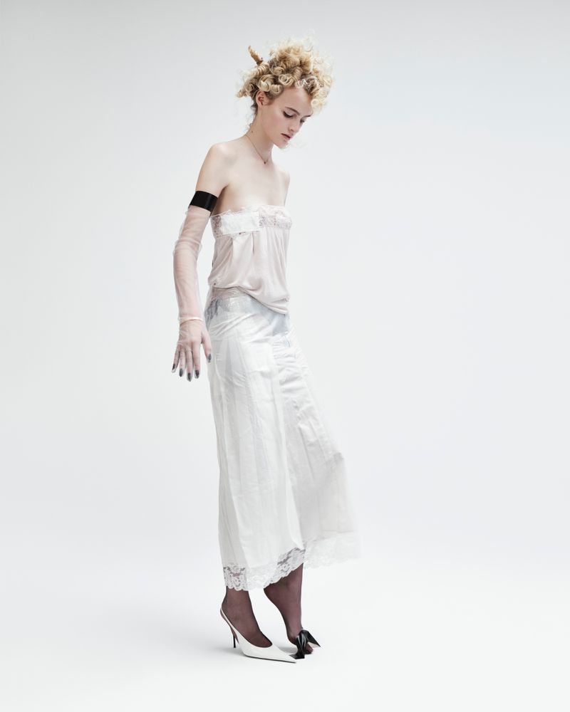 Young woman in sheer top and long slip, posing in high heels with black toe detail against a white backdrop in a fashion photograph.