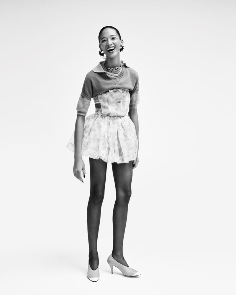 Smiling female model wearing a layered patterned outfit and pumps against a bright white background for Elle Belgium.