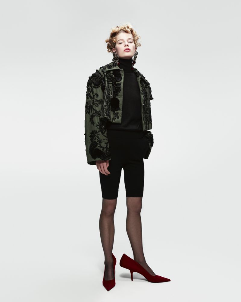 Full-length shot of a fair-skinned young woman wearing a jacket, top, and biker shorts, paired with tights and red high-heeled shoes.