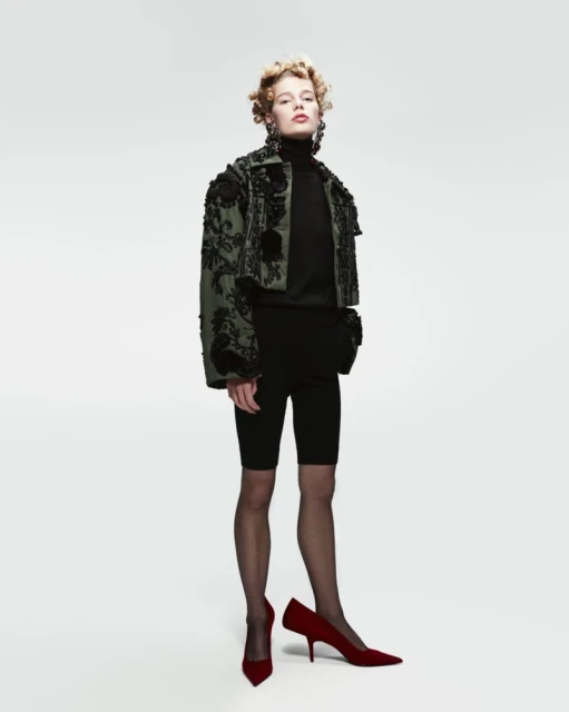Full-length shot of a fair-skinned young woman wearing a jacket, top, and biker shorts, paired with tights and red high-heeled shoes.
