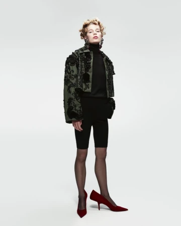 Full-length shot of a fair-skinned young woman wearing a jacket, top, and biker shorts, paired with tights and red high-heeled shoes.