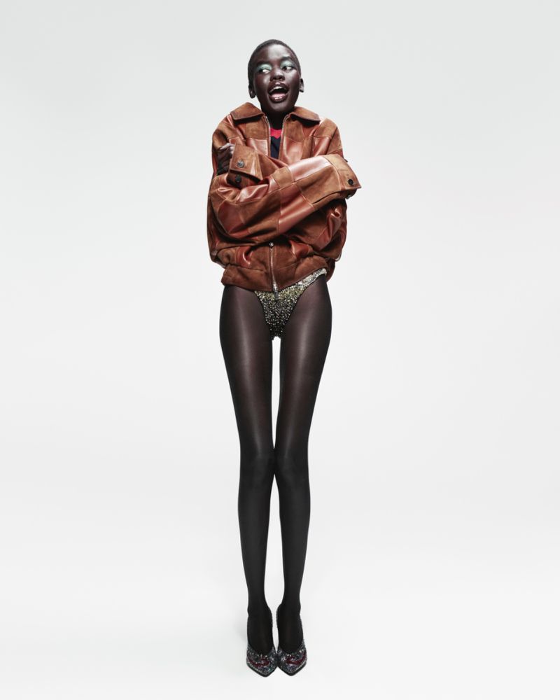 Full-length studio shot of a Black female model with an open mouth, wearing a bomber jacket, dark stockings, and heels, against a white background.