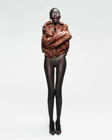 Full-length studio shot of a Black female model with an open mouth, wearing a bomber jacket, dark stockings, and heels, against a white background.