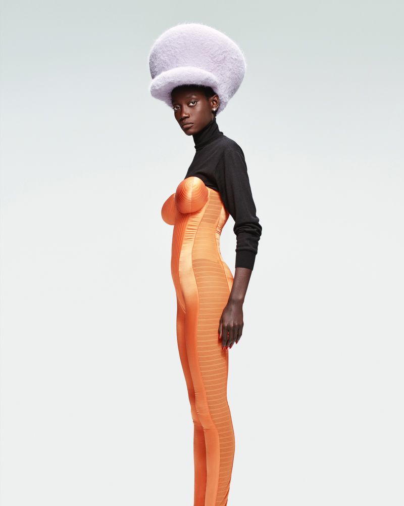 Model wearing a large furry lilac hat and orange bodysuit with black sleeves against a plain background