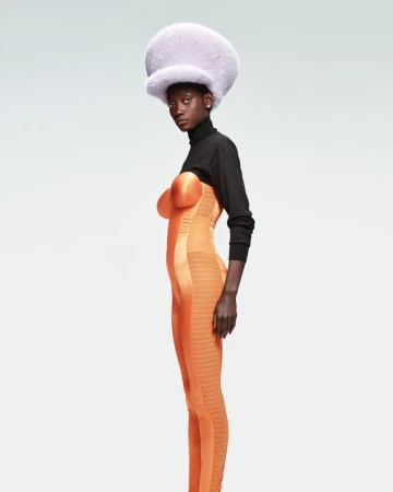 Model wearing a large furry lilac hat and orange bodysuit with black sleeves against a plain background