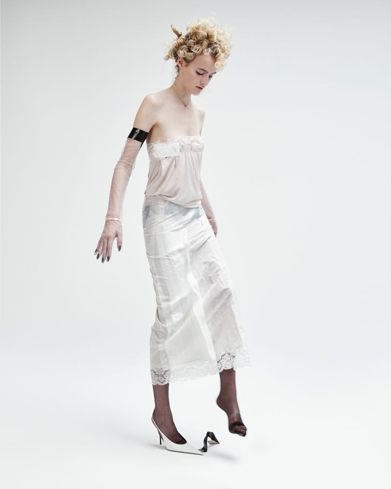 Fashion photo of a slender woman in a slip dress and heels, mid-motion against a white backdrop, striking a pose with one shoe off.