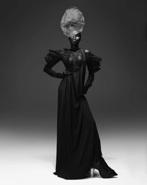 A striking full-length portrait of a dark-skinned female fashion model in an elaborate black gown and sculptural headpiece against a grey backdrop.