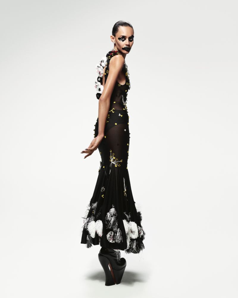 Model in a black embroidered dress with white feather details and platform shoes standing against a white backdrop.