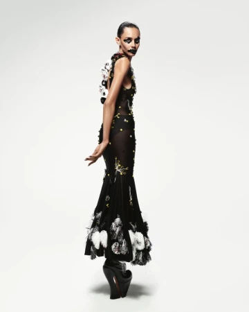 Model in a black embroidered dress with white feather details and platform shoes standing against a white backdrop.