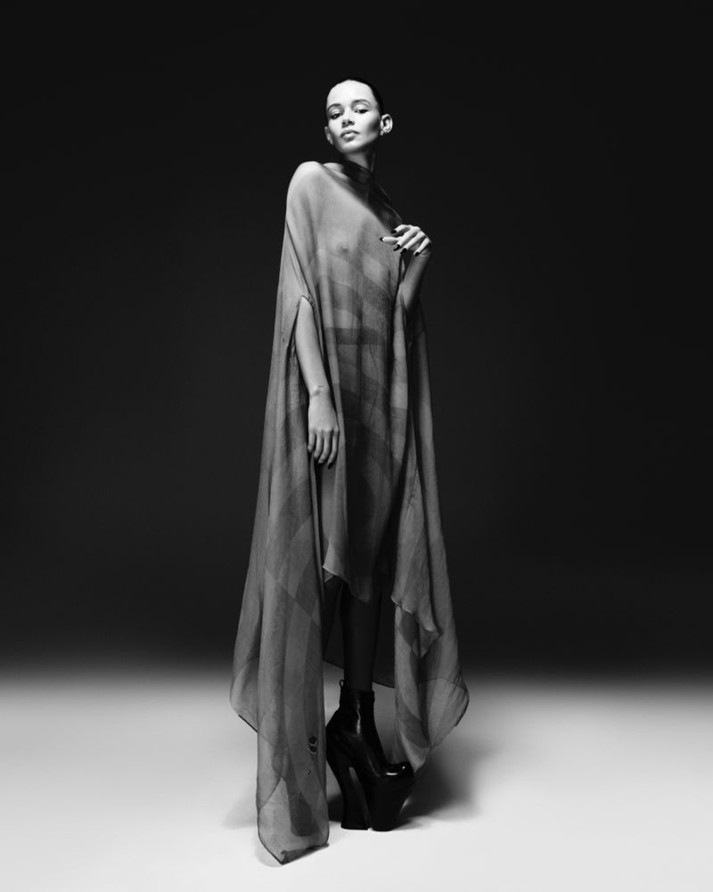 Monochrome fashion portrait of a woman wearing a sheer poncho and platform heels in a studio with neutral lighting and backdrop.