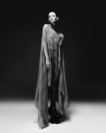 Monochrome fashion portrait of a woman wearing a sheer poncho and platform heels in a studio with neutral lighting and backdrop.