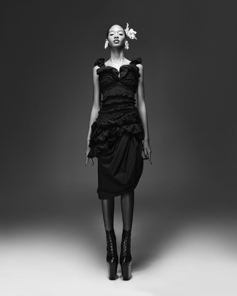 A black and white studio shot presents a slender Black female fashion model in a textured black dress and platform boots, accented with white floral accessories.