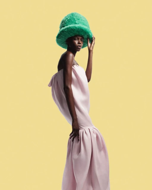 Fashionable portrait of a Black woman in a pastel pink gown and textured green hat against a yellow backdrop in a studio.