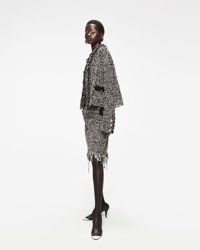 An African-American woman poses in a tweed suit with black heels against a white background, showcasing a sophisticated fashion style.