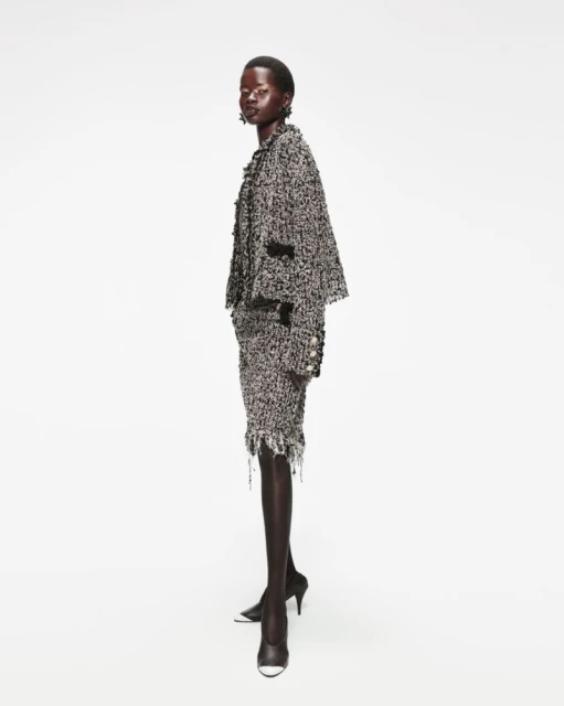 An African-American woman poses in a tweed suit with black heels against a white background, showcasing a sophisticated fashion style.