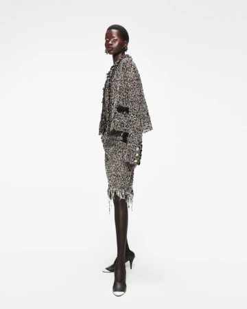 An African-American woman poses in a tweed suit with black heels against a white background, showcasing a sophisticated fashion style.