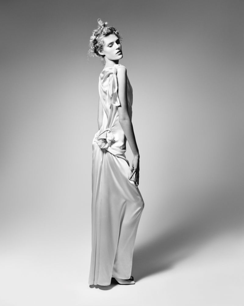 Model standing in profile wearing a pale satin gown against a neutral gradient studio background
