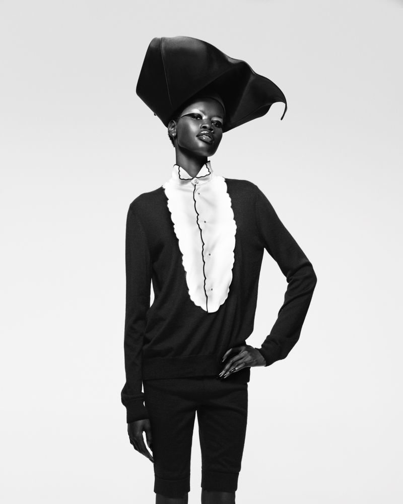 Black and white studio fashion portrait of a Black female model wearing a statement hat, blouse with ruffles, sweater, and short pants.
