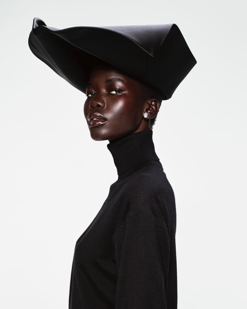 Model wearing a structured black sculptural hat and black turtleneck against a plain white background