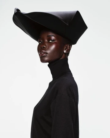 A Black model wearing an avant-garde black sculpted hat and black turtleneck sweater against a plain white background.