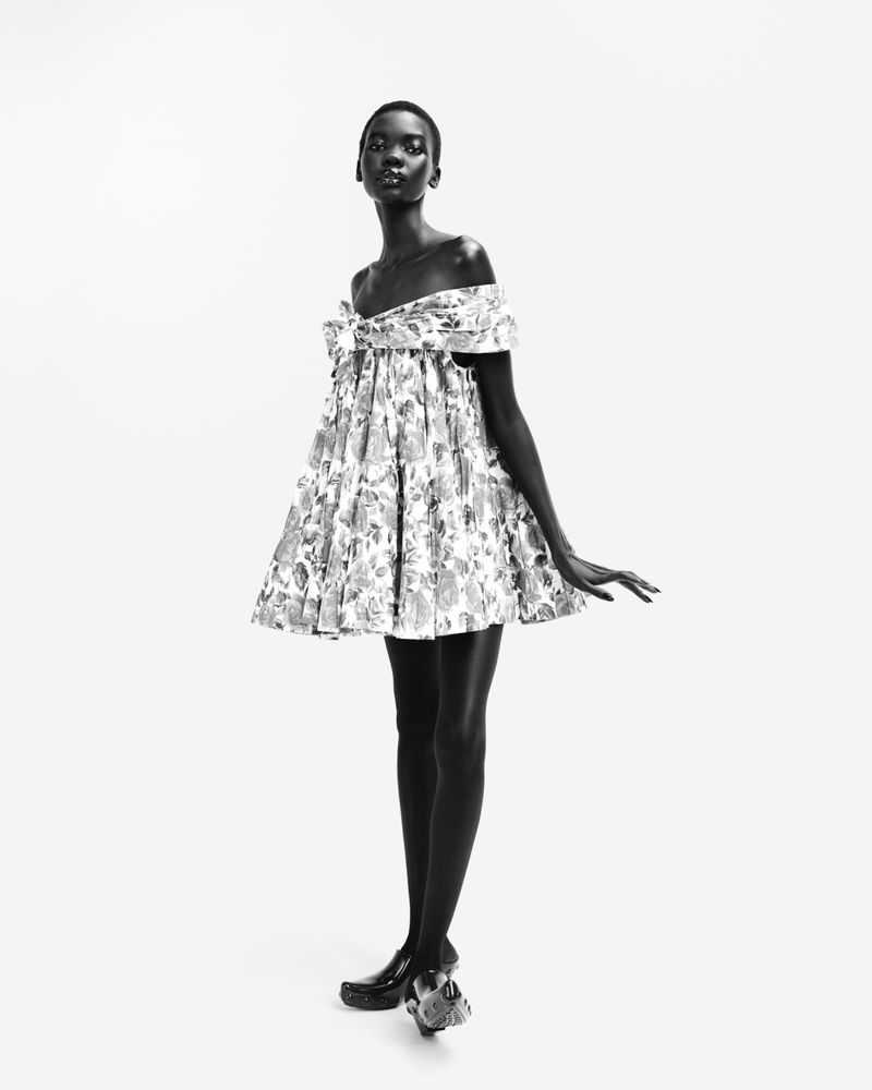 Full-length shot of a Black woman in a monochrome fashion photograph, showcasing an off-the-shoulder floral dress against white backdrop.