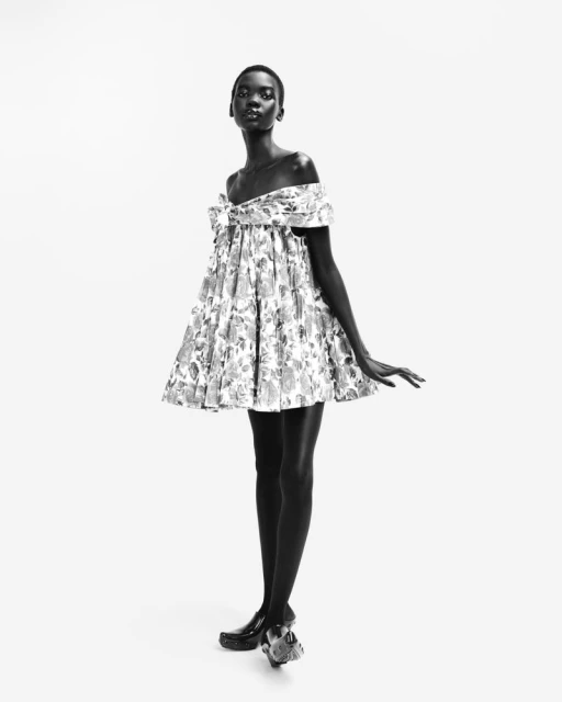 Full-length shot of a Black woman in a monochrome fashion photograph, showcasing an off-the-shoulder floral dress against white backdrop.