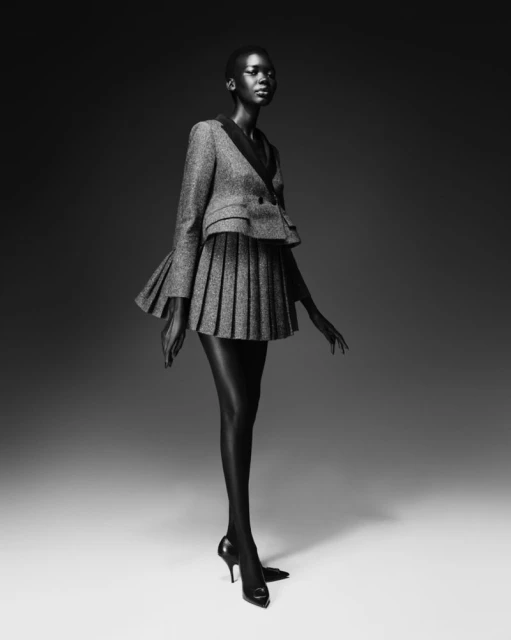 Black and white fashion shot of a Black woman in a skirt suit with a pleated skirt, black stockings and heels, posing against a gradient gray backdrop.