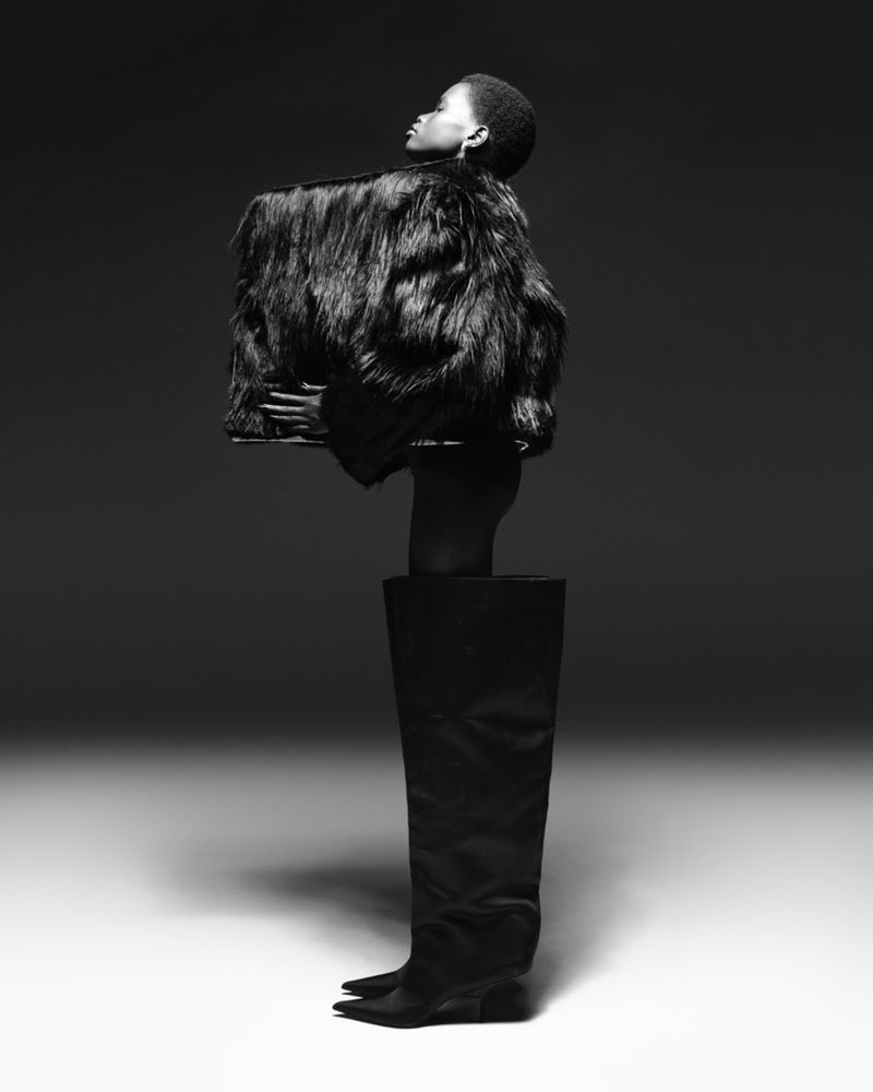 Monochrome fashion portrait of a Black woman in a fur coat and thigh-high leather boot against a smooth gradient backdrop.