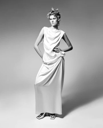Model standing in a flowing white dress against a neutral grey studio background for Elle Belgium.