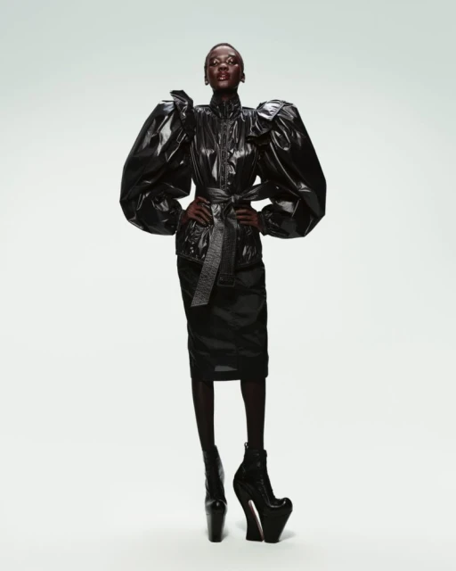 Fashion model in a studio setting wearing a shiny black jacket with large puffy sleeves, a matching skirt, and high-platform boots.