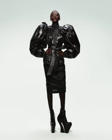 Fashion model in a studio setting wearing a shiny black jacket with large puffy sleeves, a matching skirt, and high-platform boots.