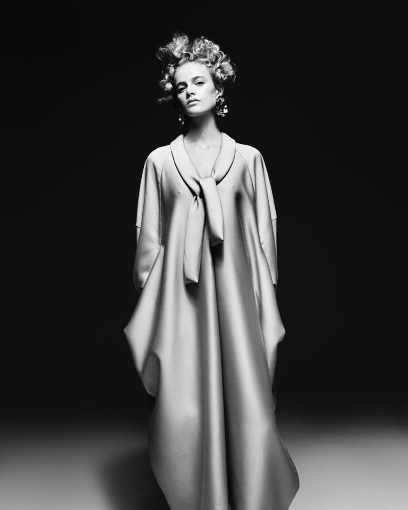 Black and white fashion portrait of a blonde woman with an elaborate updo, wearing a light-colored dress with a tie at the neckline.