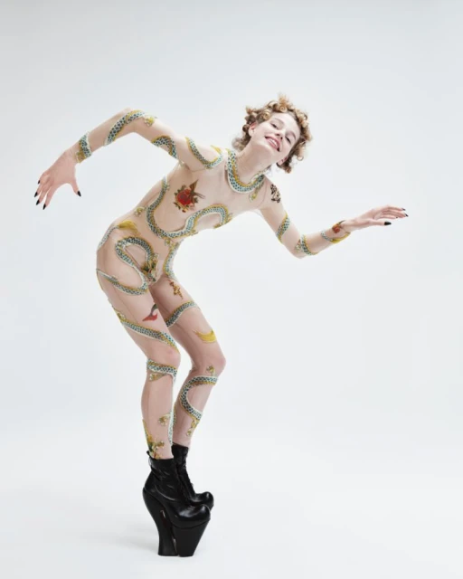 A woman with curly hair and a bodysuit with snakes is posed in front of a white background. She is also wearing platform shoes.