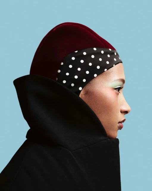 Profile portrait of woman with light skin wearing a red velvet hat with black and white polka dot band and a black hooded coat.
