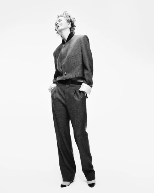 Full-length black and white studio shot of a smiling adult female model with curly hair wearing a blazer and wide-legged pants.