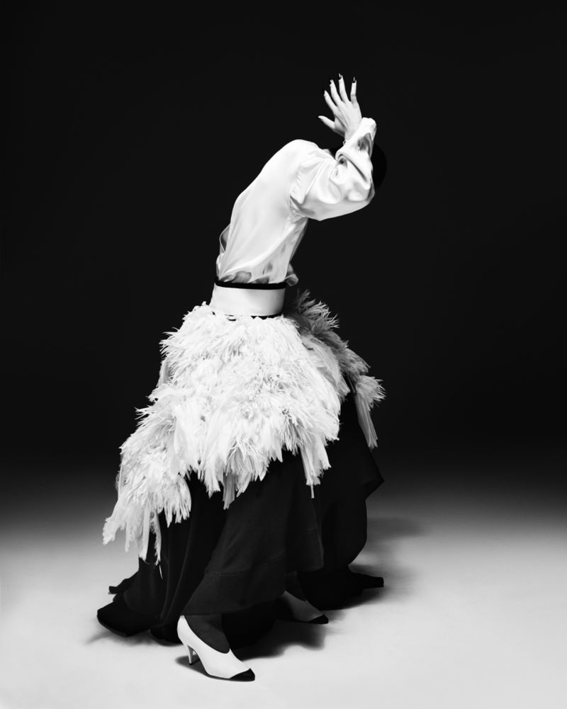 Striking black and white fashion portrait of a person with raised hands, wearing a white shirt and feathery black and white skirt.