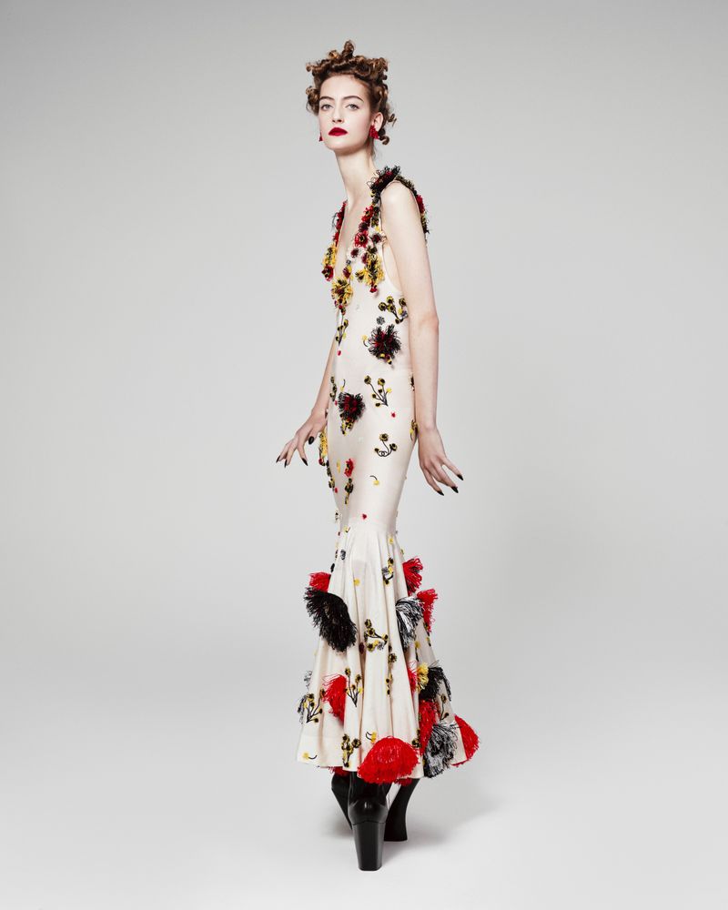 A striking, full-length studio shot of a poised model showcasing a cream-colored gown with bold red and black floral accents and black boots.