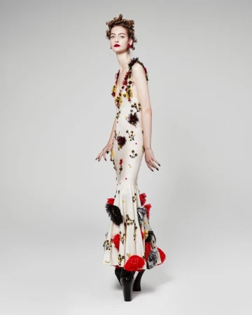 A striking, full-length studio shot of a poised model showcasing a cream-colored gown with bold red and black floral accents and black boots.