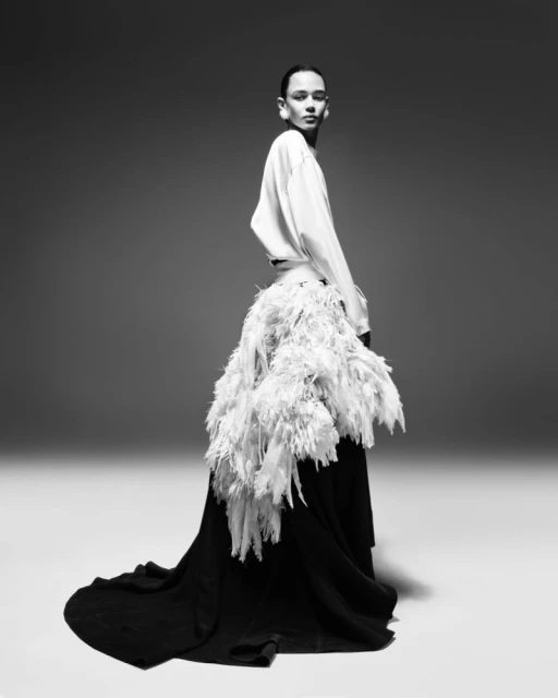 Black and white fashion shot of a person with long dark hair wearing a flowing white shirt and feathered black dress posing in studio.