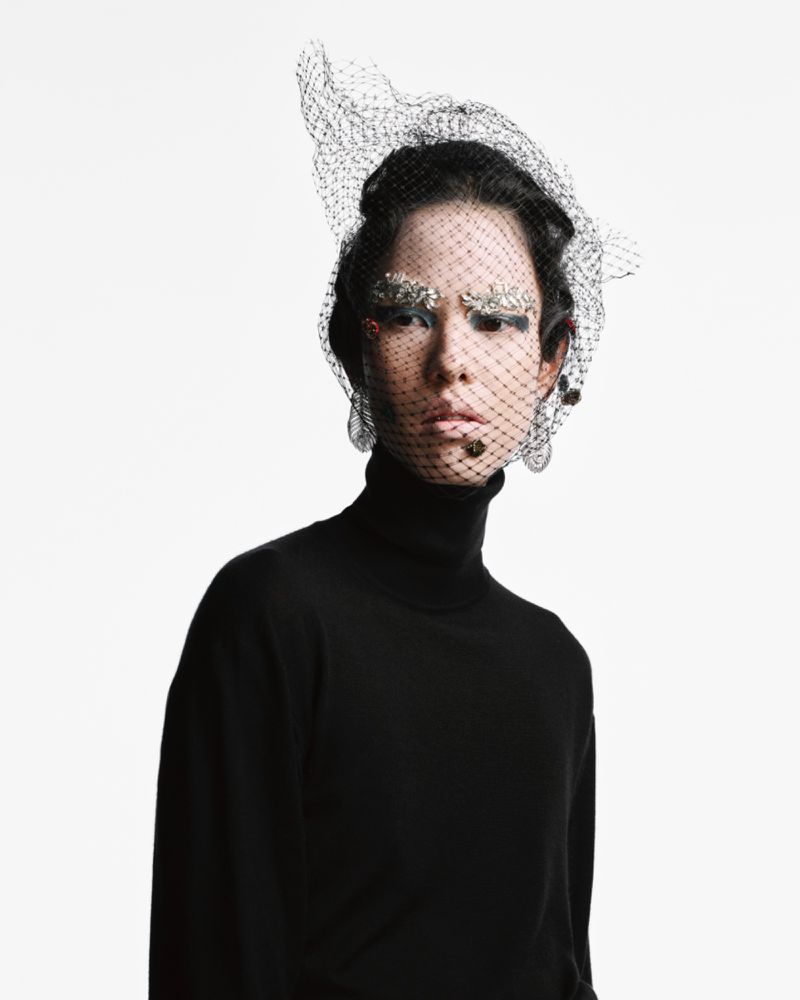 Close-up portrait of a model wearing a black turtleneck sweater and elaborate net headpiece with embellished eyebrow makeup against white.