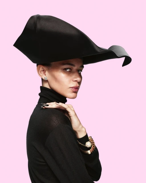 Elegant woman in black turtleneck and statement hat against pink backdrop, accessorized with gold bracelet and nail art.