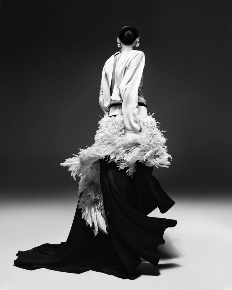 Back view of adult female fashion model in feathered collar and gown, studio shot in black and white. High fashion editorial concept.