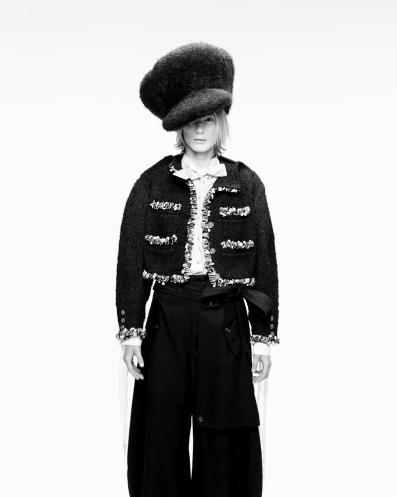 Full-length black and white portrait of an adult woman in a dark hat, textured jacket with light trim, white blouse, and wide-leg pants.