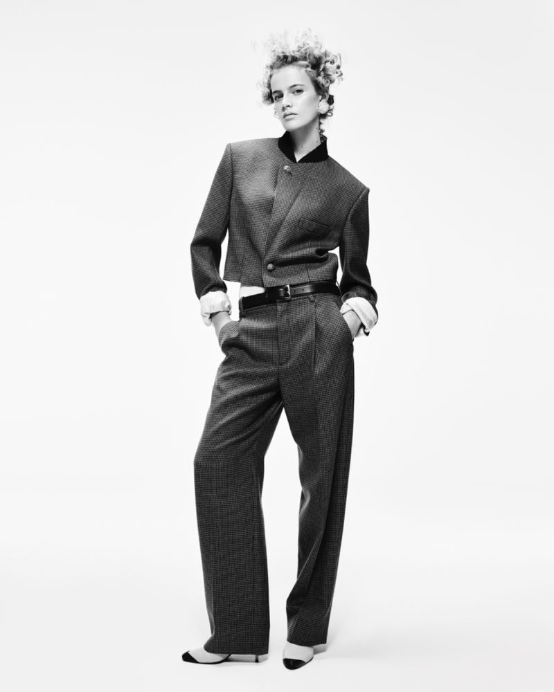 Blach-and-white full shot of a woman in a tailored suit with her hands in her pockets, striking a confident pose against a plain background.